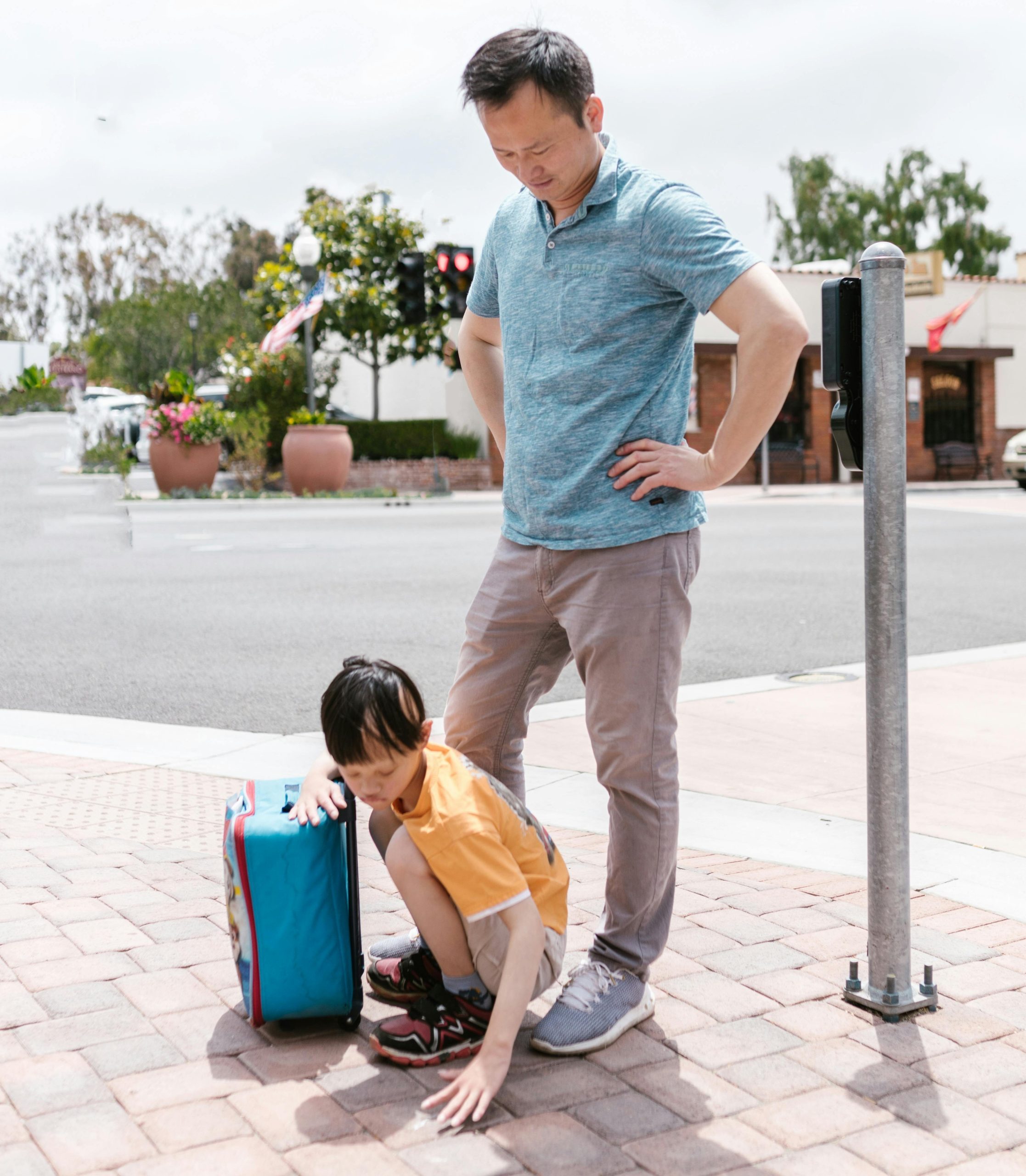 Preparing for Summer Travel with ABA Therapy Man standing with hands on hips watches two kids examining a blue rolling suitcase on a brick sidewalk outdoors.