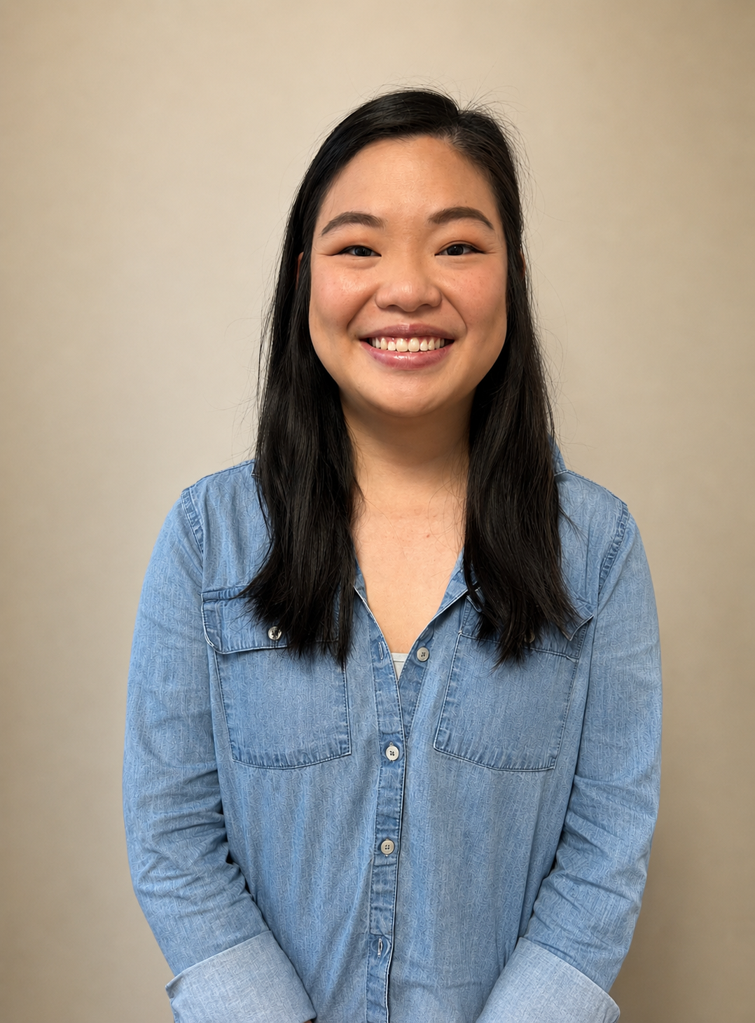 Portrait of a Smiling Woman with Long Black Hair Wearing a Blue Denim Shirt Against a Beige Backdrop.